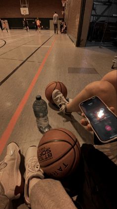 two basketballs are laying on the floor while someone is holding a cell phone in their hand