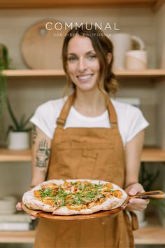 a woman in an apron holding a pizza on a wooden platter with toppings