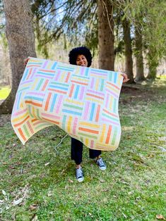 a woman is holding up a blanket in the grass with trees behind her and smiling at the camera