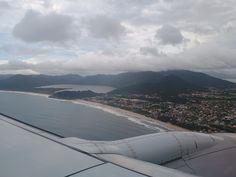 the wing of an airplane as it flies over a beach and ocean with mountains in the background