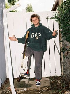 a young man standing in front of a white fence with his arms out and holding a guitar