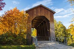 a wooden covered bridge in the fall with trees and leaves around it, on a sunny day