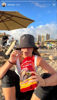 a woman sitting on the beach holding a bag of chips in her hand and an umbrella over her head