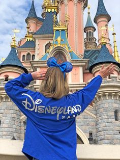 a woman standing in front of a castle wearing a blue shirt with disney land on it