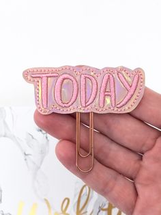 a hand holding a pink pin with the word today on it and a gold clip