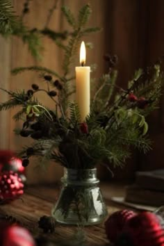 a candle is lit in a glass vase filled with greenery and pine cones on a table