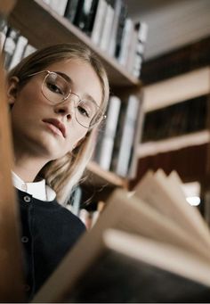 a woman wearing glasses reading a book in front of a bookshelf full of books