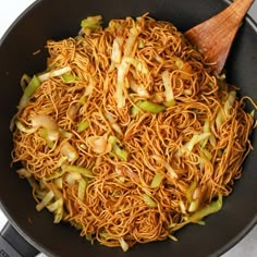 a wok filled with noodles and vegetables on top of a white table cloth next to a wooden spoon