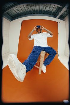 a man sitting on top of a wooden chair in front of an orange wall with his hands behind his head