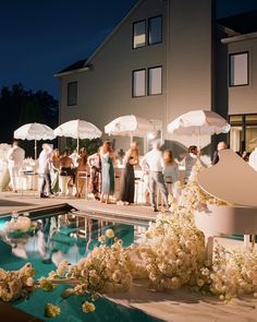 a group of people standing next to a pool with white umbrellas over it and flowers on the ground