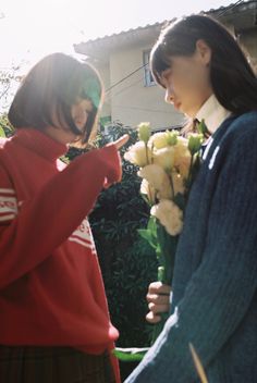 two young women standing next to each other in front of a house with flowers on the ground