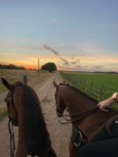 two people riding horses on a dirt road near a grassy field and fence at sunset