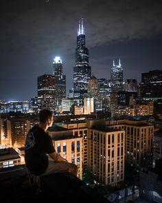 a man sitting on top of a roof looking out at the city lights in the distance