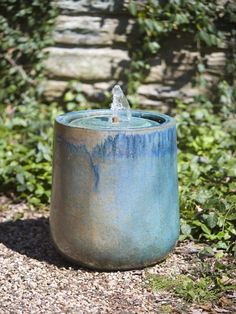 a blue pot sitting on top of a dirt ground next to bushes and trees with water coming out of it