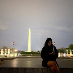 a woman sitting on a ledge looking at her cell phone in front of the washington monument