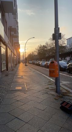 the sun is setting on an empty city street with cars parked along the side walk