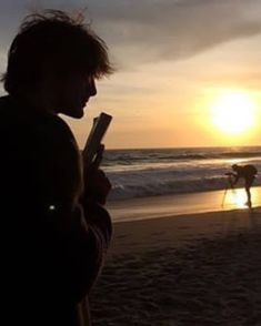 a man standing on top of a beach next to the ocean holding a cell phone