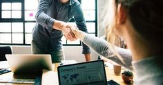 two people shaking hands in front of a laptop computer on a desk with other people