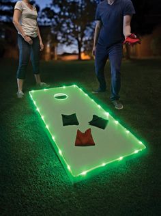 a man and woman standing next to a lighted cornhole game in the grass at night