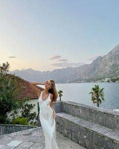 a woman standing on top of a stone walkway next to a body of water with mountains in the background