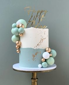 a blue and white cake with gold decorations on top is sitting on a stand in front of a gray wall