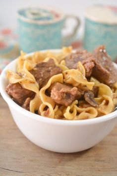 a white bowl filled with beef and noodles on top of a wooden table next to two cups