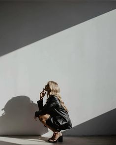 a woman sitting on the ground talking on her cell phone in front of a white wall