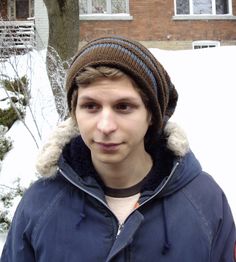 a young man wearing a beanie standing in front of a house with snow on the ground