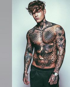 a man with tattoos standing in front of a white wall and looking at the camera