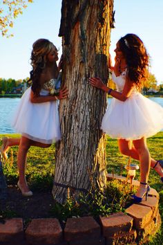 two girls in white dresses standing next to a tree and one girl is leaning against the tree