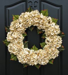 a white wreath hanging on the front door of a black door with green leaves and flowers