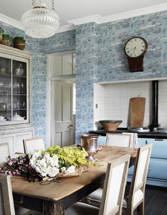 a dining room table with flowers on it in front of a wallpapered kitchen