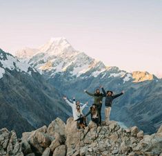 four people standing on top of a mountain with their arms in the air and mountains behind them