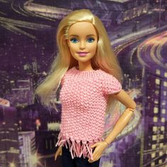 a barbie doll with blonde hair and blue eyes wearing a pink knitted sweater in front of a cityscape backdrop