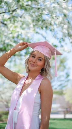 a woman wearing a pink graduation cap and gown standing under a tree with her hands on her head