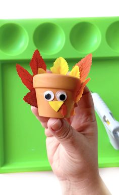 a hand holding a clay pot with a fake turkey in it's beak and leaves on its head