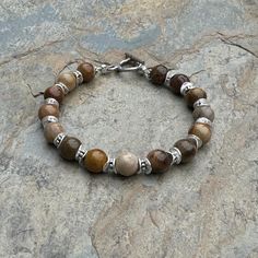 This petrified wood bracelet embraces the mystique of nature with a gemstone that has transformed over millions of years. Each round is separated with sterling silver spacer beads. The intricate, natural patterns within the petrified wood add a touch of ancient charm. Strung on sturdy, high quality beading wire and closes with a sterling Hill Tribe silver  toggle. Please message me if you need a different size or clasp. 7.5 inch bracelet Petrified wood rounds alone: 8mm  Your jewelry will arrive in a cotton lined kraft box within a padded postal envelope. I use USPS first class mail. This usually takes 3 to 5 days to ship. International orders outside of the U.S. will take 8 to 15 days to ship. To see more from East Village Jewelry~ http://eastvillagejewelry.etsy.com All orders ship out wi Artisan Brown Bracelets With Gemstone Beads, Artisan Brown Bracelet With Gemstone Beads, Earthy Brown Jasper Beaded Bracelets, Earthy Jasper Gemstone Bead Bracelets, Earthy Jasper Gemstone Beads Bracelet, Earthy Brown Bracelets With Gemstone Beads, Rustic Brown Bracelets With Natural Stones, Earthy Brown Gemstone Beads Bracelets, Brown Jasper Round Bead Bracelets