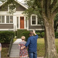 a man and woman standing in front of a house waving to the sky with their hands up