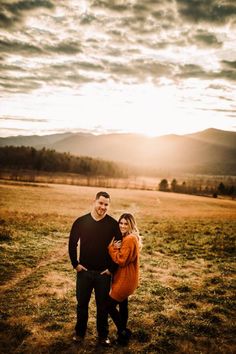 a man and woman standing in an open field with mountains in the background at sunset