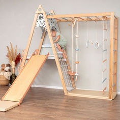 a child playing on a wooden slide next to a wall with toys hanging from it