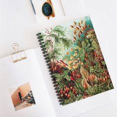 an open spiral notebook on top of a desk with pictures and pens next to it