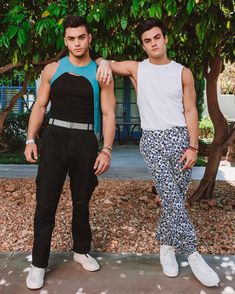 two young men standing next to each other in front of some trees and bushes with their arms around one another