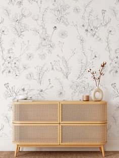 a white wall with flowers on it next to a wooden dresser and vase in the foreground