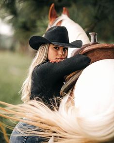 a woman wearing a cowboy hat and black sweater sitting on a white horse with long blonde hair