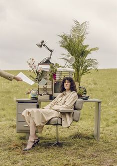 a woman sitting at a desk in the middle of a field with plants and books on it