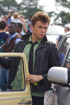 a young man standing next to a parked car in front of a group of people