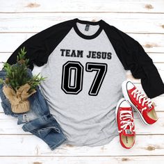 This Team Jesus Jersey Shirt is the perfect Christian shirt for any sports fan! It's a sporty stylish way to share your faith and heart for Jesus with anyone! Plus it's super soft and comfy. Whether your heading to your favorite sports game, running errands or a weekend get away, this cute Team Jesus shirt is a comfy casual Jesus shirt for any occasion. A stylish spin on the classic baseball raglan. The combed cotton blend makes it super soft, comfortable, and lightweight. You don’t even have to Black Tops With Lettering For Sports Events, Black Collegiate Tops With Lettering, Team Spirit Tops With Lettering For Game Day, Casual T-shirt With Lettering For Game Day, Casual Athletic Heather T-shirt For Game Day, Casual Gray Top For Game Day, Game Day Athletic Heather Casual T-shirt, Varsity Sports Top With Lettering, Black Sporty Tops With Lettering