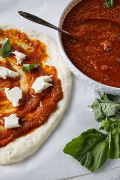 a pizza sitting on top of a table next to a bowl of sauce and basil