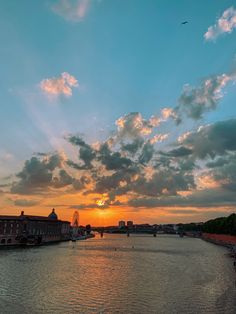 the sun is setting over some water with buildings in the background and clouds in the sky