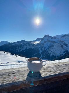 a cup of coffee sitting on top of a wooden table next to snow covered mountains